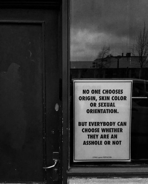 Derrière une vitre à la droite d’une porte, une affiche sur laquelle est écrit : No One chooses origin, Skin color, or sexual orientation. But everybody can choose whether they are an asshole are not.