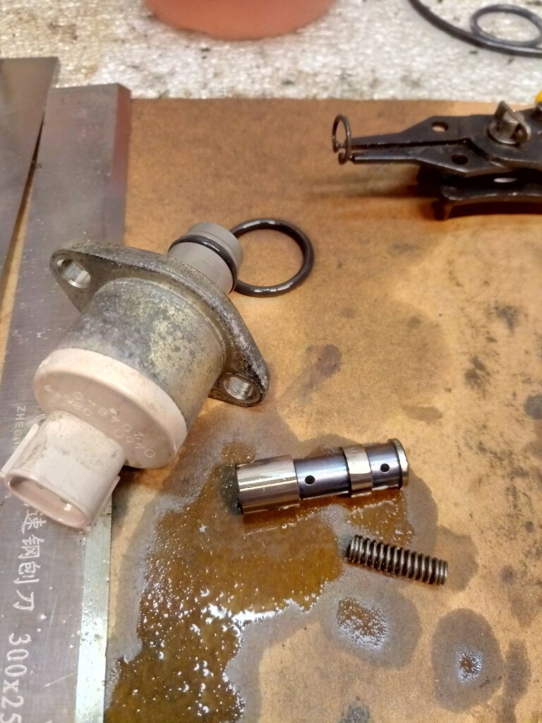 Fuel metering valve from a diesel injector pump. The valve is taken apart. A spring, sliding piston with holes and o-ring are strewn around. A circlip plier has an inner circlip impaled on it.