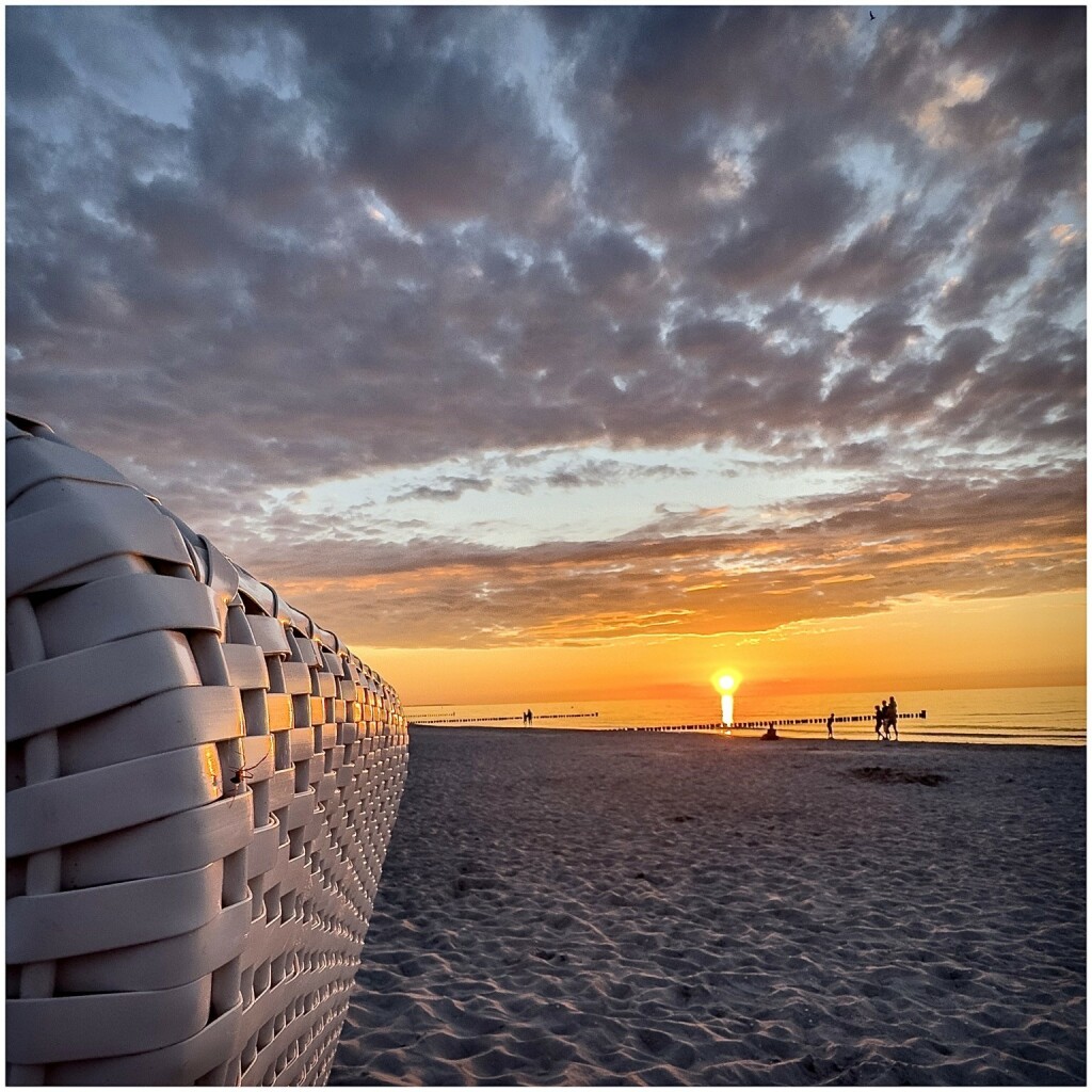 A beachfront sunset with a partly cloudy sky, people on the shore, and a wicker beach chair in the foreground.