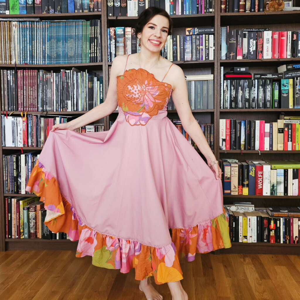 Marina vor einem Bücherregal in einem rosa Kleid mit breitem Volant und einer Rosenapplikation in Orange, lila, rosa und hellgrün. 