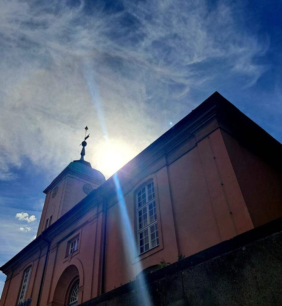 Farbfoto: Von unten nach oben fotografiertes großes, bräunliches Gebäude mit einem Turm, auf dem man eine Wetterfahne sieht. Oben blauer Himmel mir Federwolken. Direkt über dem Gebäude die Sonne, die einen dicken  blauen Strahl nach unten schickt.