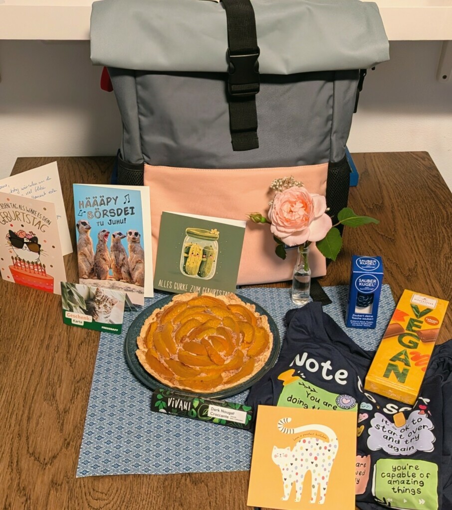 ein Tisch mit Geschenken: Geburtstagskarten, ein kleiner Aprikosenkuchen, diverse kleine Geschenke und Schokolade, ein Rucksack in Grau, Schwarz und Rosa und eine rosa Rose in einer kleinen Glasvase.