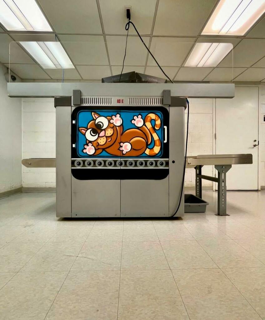 Streetartwall. On the outside of a large suitcase scanner machine in the
World Trade Center, a small mural with a cat was sprayed/painted. The large metal machine takes up an entire bare, tiled room. The little cartoon cat in the middle of the machine adds a little color and fun. She is being scanned and is lying in a suitcase that is rolling over a conveyor belt. It has its paws pressed against the glass (the suitcase) and is looking at the viewer with wide eyes.