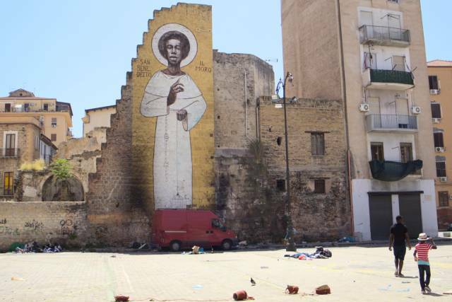 A somewhat shabby square with a large graffiti on a wall depicting St Benedict the Moor. Palermo, Sicily