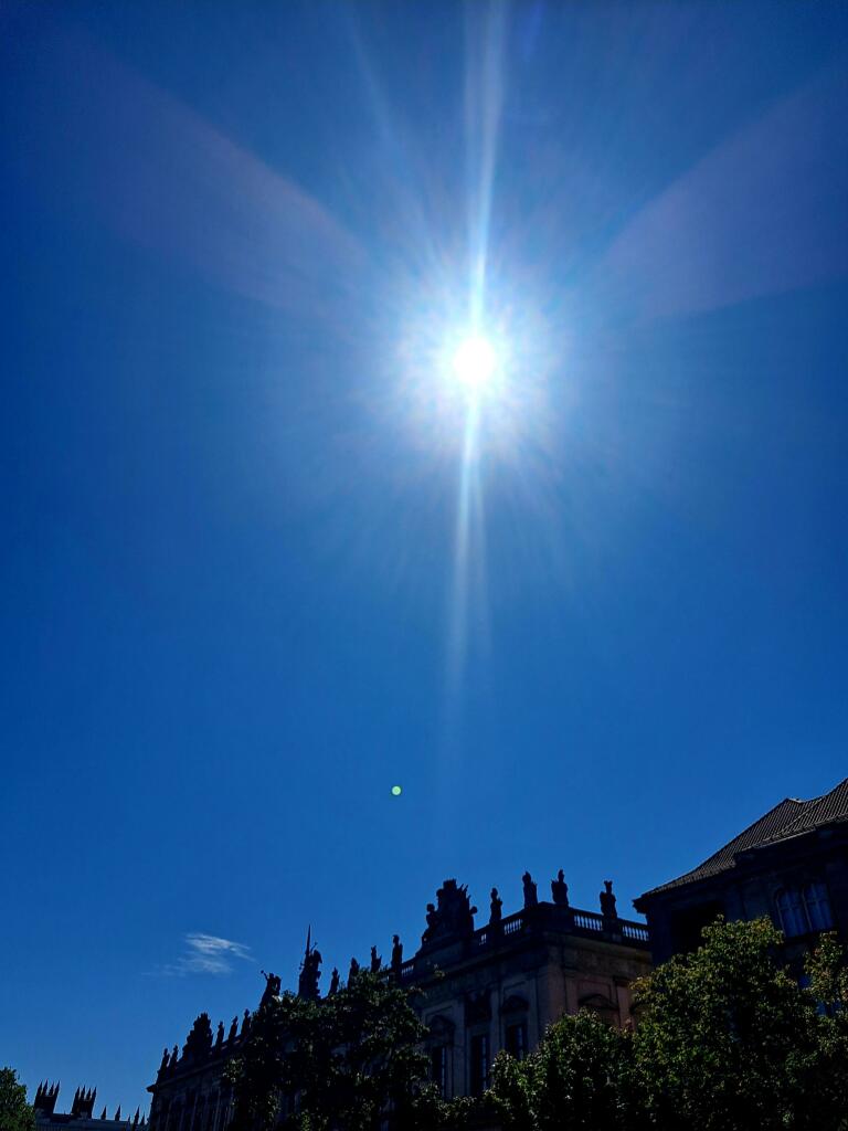 Farbfoto: Im unteren Teil des Bildes sieht man den oberen Teil eines Gebäudes, ansonsten nur tiefblauen Himmel und die Sonne mit ihren Strahlen.