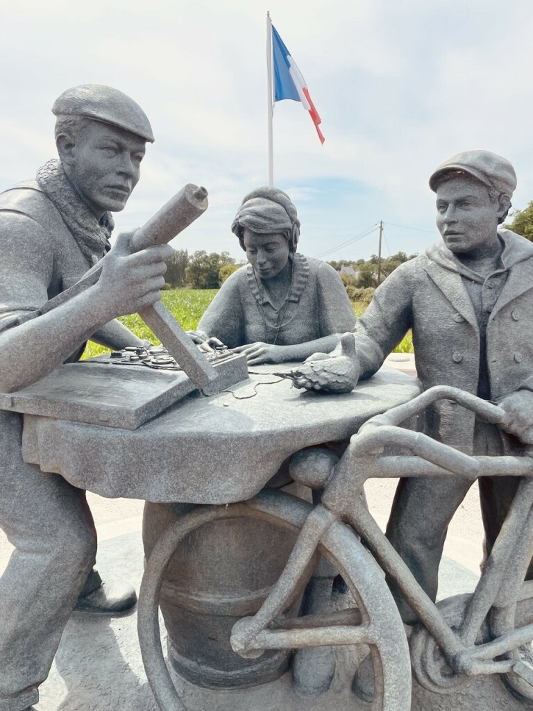 Foto eines figürliches Denkmals aus Beton, mit denen im Post benannten Personen. Die Aufgaben werden symbolisiert über Utensilien, welche den einzelnen Personen beigefügt werden: Waffe (Guerilla), Rundfunkempfänger mit Kopfhörern (?, Spionage), Brieftaube und Fahrrad (Untergrund). Im Hintergrund ländliche Landschaft und ein Fahnenmast mit französischer Flagge.