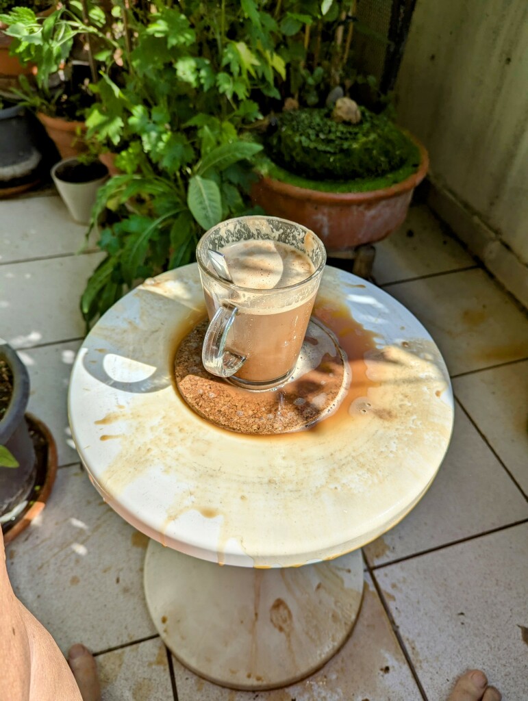 Auf einem ehemals weissen Kunststoffhocker steht eine halbleere Glastasse mit Cappuccino.
Der Rest vom Cappuccino ist auf Hocker, Fußboden und meinen Zehen verteilt.