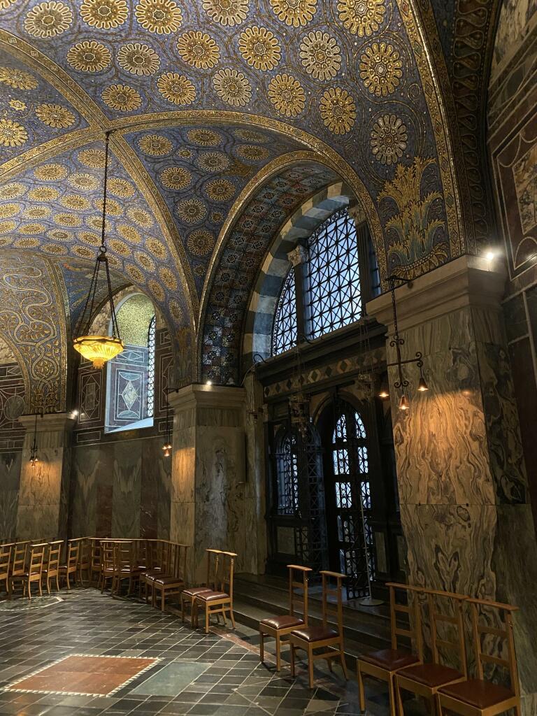 Ornate interior of a historical building with a richly decorated, arched ceiling featuring intricate gold and blue patterns. Rows of wooden chairs line the floor, and ornate windows are on the far wall.