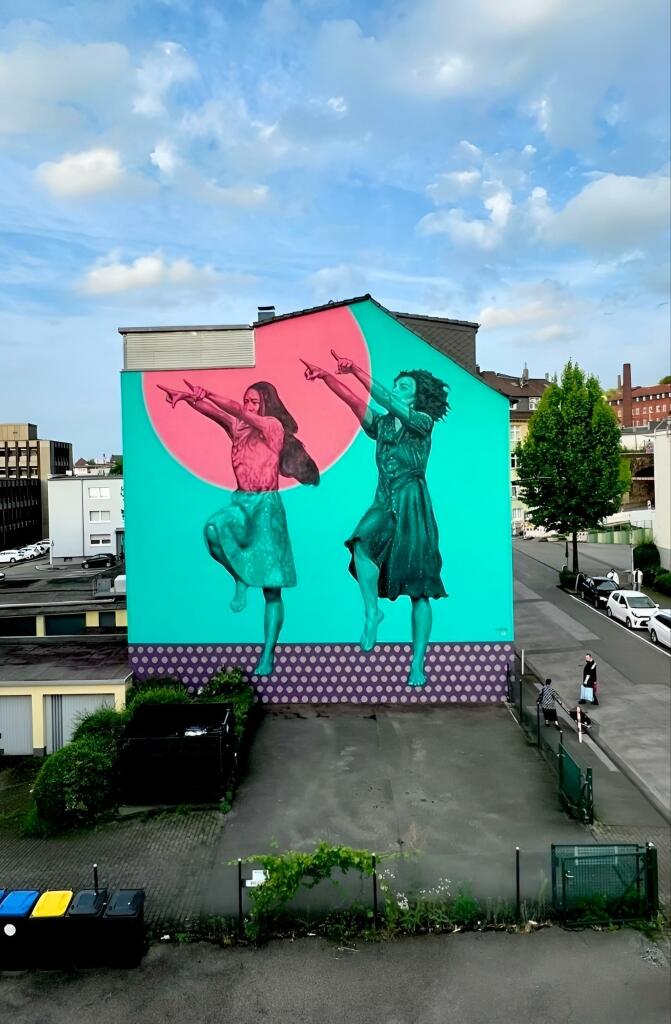 Streetarteall. A huge mural with two dancing young women was sprayed/painted on the outside wall of a four-storey building. The background is turquoise, the figures are drawn in black.  It shows two young women in summer dresses, both arms pointing upwards towards a red sun and jumping over a purple dotted floor. A wonderful homage to modern dance theater.
Info: The second mural of a total of ten planned murals as a tribute to the great director/choreographer Pina Bausch (1940 - 2009) as part of the "Pina Bausch Gallery - Wuppertal dances" project. "Palermo Palermo" is the title of the dance piece that the artist dedicated to the Sicilian capital and which premiered in Wuppertal in 1989. For their research, the dance company lived in the city for several weeks, got to know life in the city, met lots of people and then realized the piece.