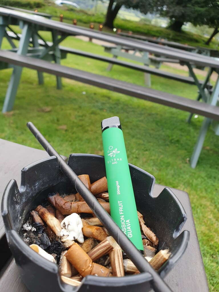 A green Elfbar disposable vape, labelled 'Passion Fruit Guava' (20mg/ml), sits in an ashtray filled with cigarette butts, placed on an outdoor table with picnic benches in the background.