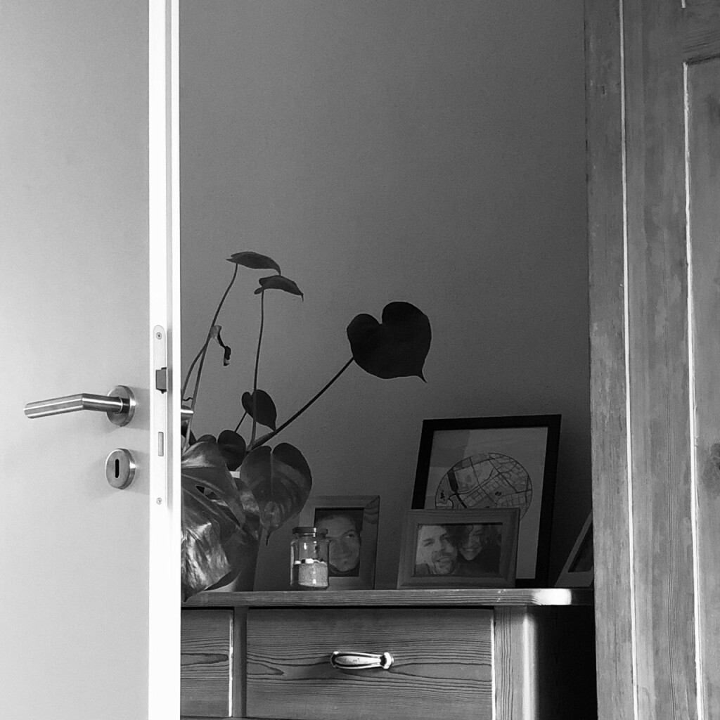 Black and white photo taken in a bedroom. White door leaf with steel door handle on the left, old wooden closet on the right, white wall and a wooden dresser in between. On the dresser there are a monstera in a barely visible white pot, a small jam jar filled with beach sand and seashells as well as pictures in wooden frames. One of them shows a happy young couple.