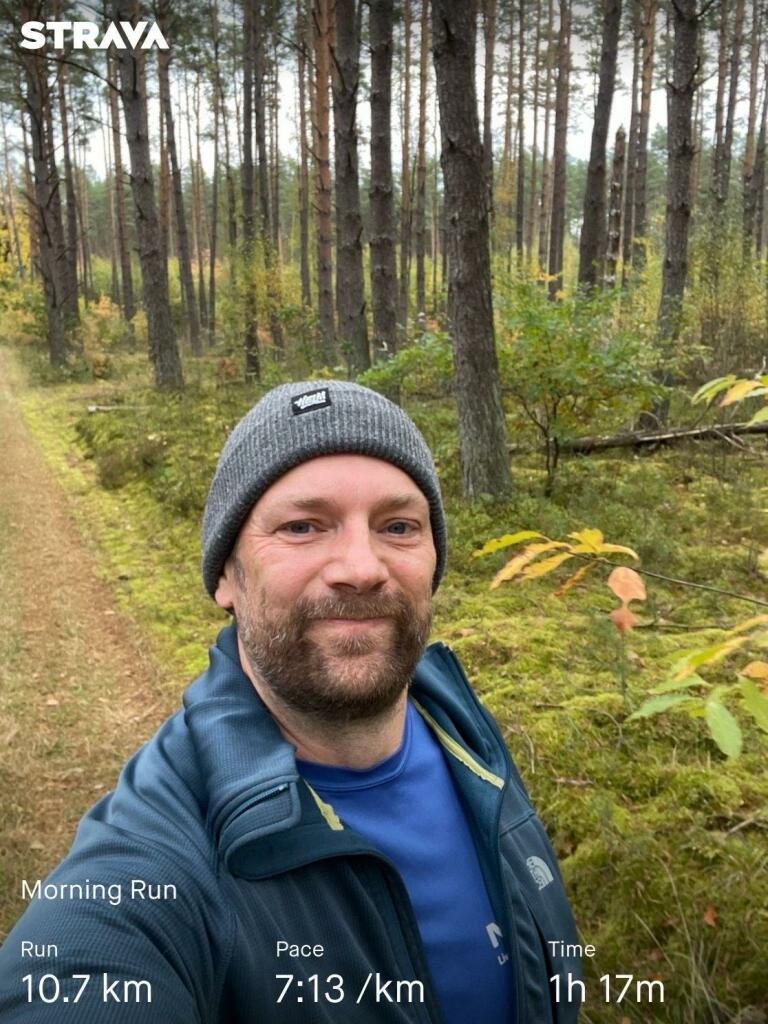 Ein Mann lächelt, während er während eines morgendlichen Laufs in einem Waldgebiet ein Selfie macht. Er trägt eine Mütze und eine Sportjacke. Zu den angezeigten Informationen gehören die Laufstrecke (10,7 km), das Tempo (7:13/km) und die Gesamtzeit
