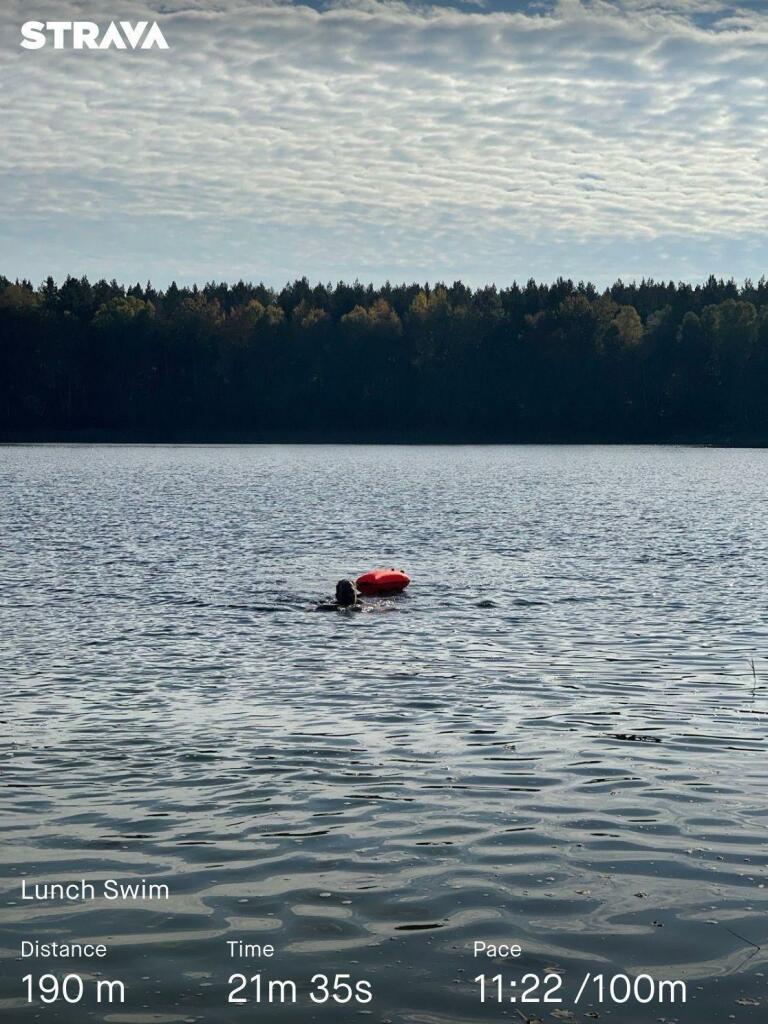 A person swims in a lake with a floating device nearby. The scene features calm water under a cloudy sky, with trees lining the background. Overlayed text indicates a distance of 190 meters, a time of 21 minutes and 35 seconds,