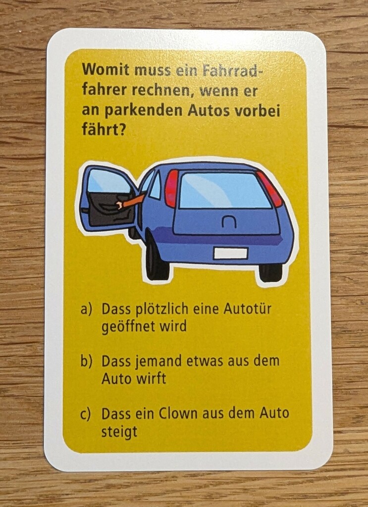 Eine gelbe Karte mit weißem Rand aus einem Fahrradquiz für Kinder, mit dem Bild eines blauen Auto und einer geöffneten Fahrertür mit Hand am Griff.

Frage: Womit muss ein Fahrradfahrer rechnen, wenn er an parkenden Autos vorbeifährt?

a) Dass plötzlich eine Autotür geöffnet wird
b) Dass jemand etwas aus dem Auto wirft
c) Dass ein Clown aus dem Auto aussteigt