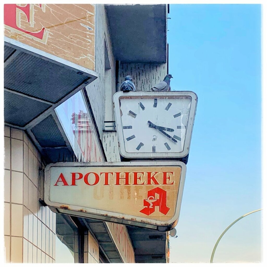 Eine Uhr und ein Schild für eine "Apotheke" (Apotheke) sind auf einem Gebäude montiert. Zwei Tauben ruhen auf der Uhr. Der Hintergrund zeigt einen klaren blauen Himmel.