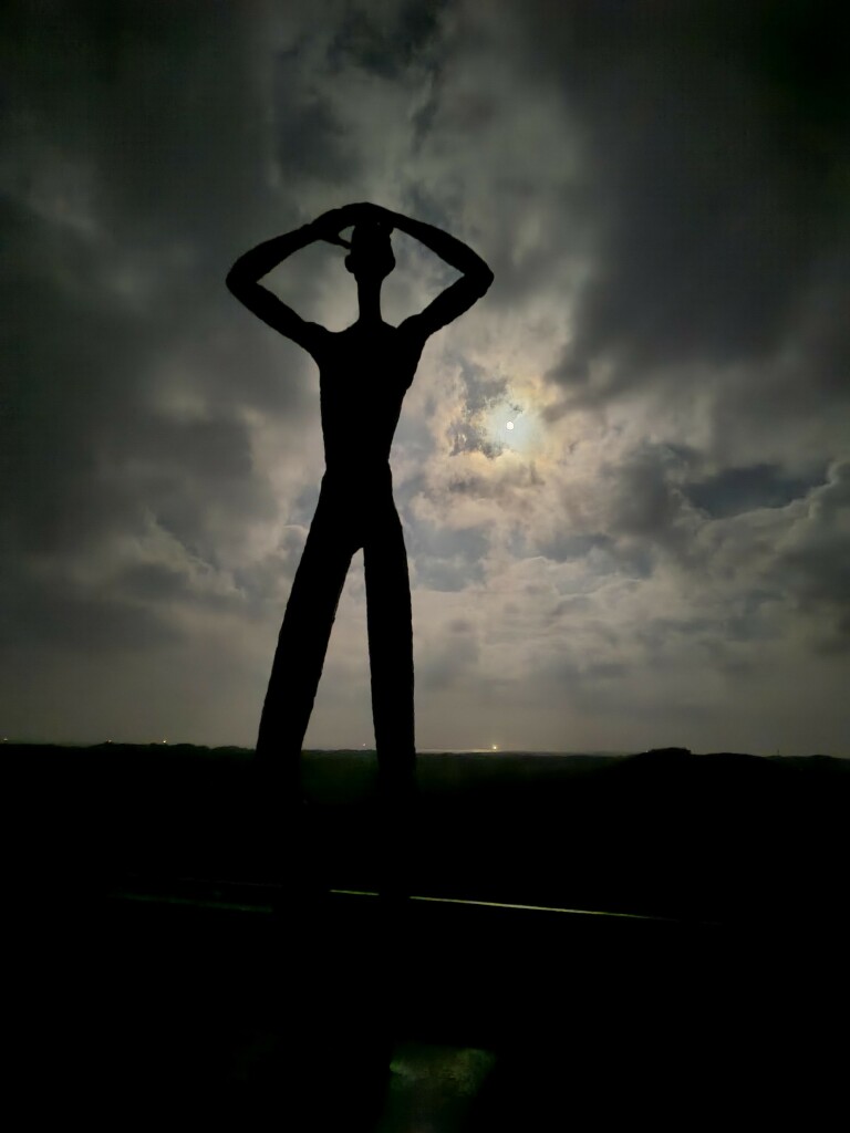 Statue an erhöhtem Platz (Utkieker), die sich die Hände an den Kopf hält (wie wenn man gegen Sonne weit gucken will). Nachts mit hellen, vom Mond angestrahlten weißen Wolken.