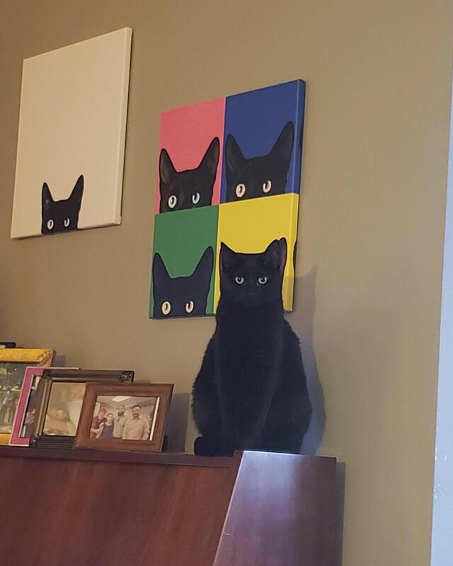 A cat standing in front of a painting that shows only the top half of faces of cats. The cat is blending in with the background painting.