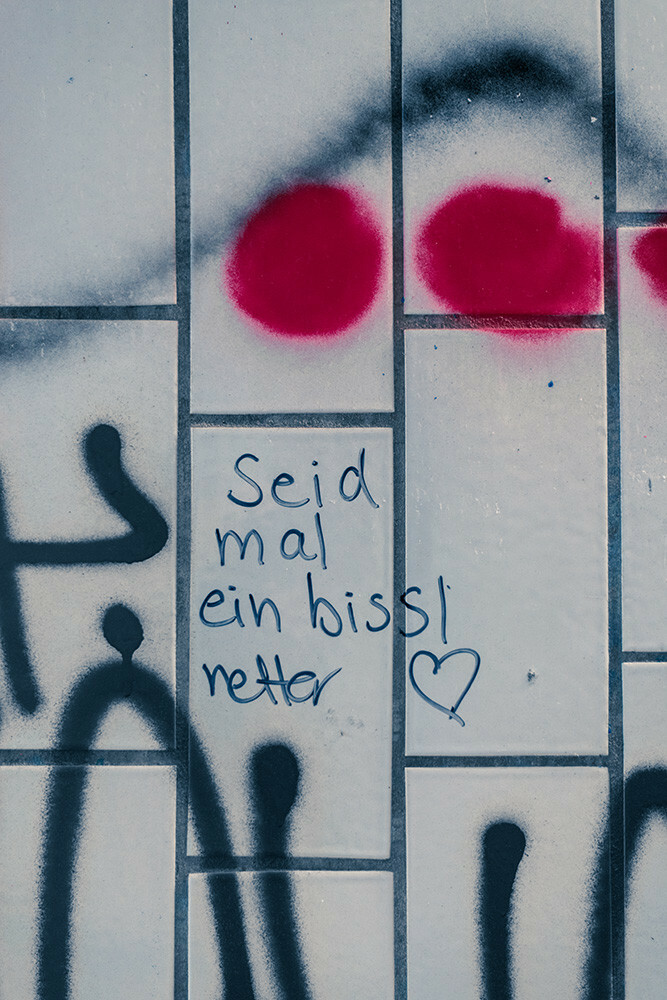 An der weiß gefliesten Wand einer Fußgänger-Unterführung steht zwischen verschiedenen Graffitis "Seid mal ein bissl netter" mit einem schwarzen Marker geschrieben und einem Herz dahinter.