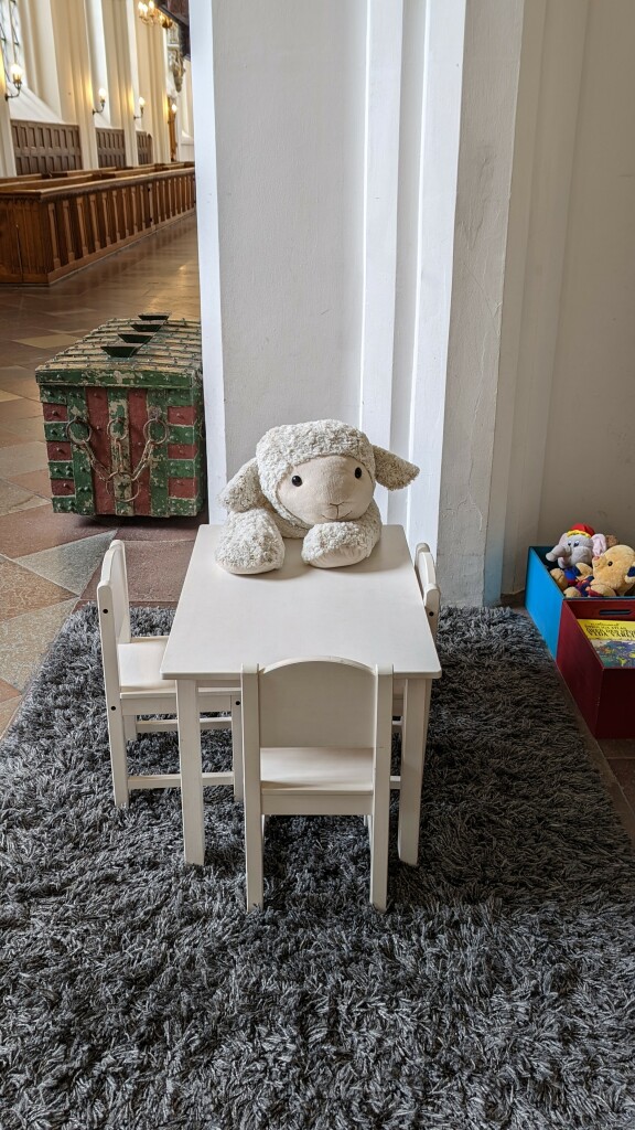 Ein Plüsch-Lämmchen sitzt an einem sehr kleinen Tisch für Kinder in einer Spielecke in einer Kirche, im Hintergrund steht eine große Truhe, es sind weitere Spielsachen zu erkennen sowie Sitzbänke 