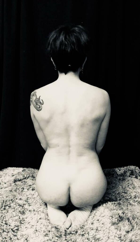 A nude person is kneeling with their back to the camera. They have short hair and a tattoo of an octopus on their shoulder. The background is dark, and the image has a black-and-white filter. The person is seated on a textured surface