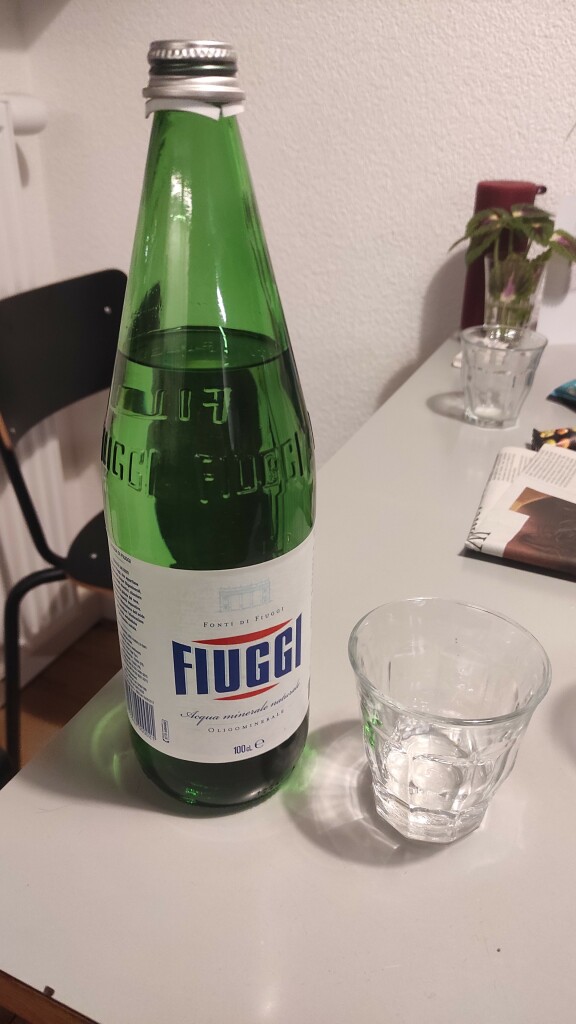 Eine italienische grüne Wasserflasche der Marke Fiuggi steht auf einem Küchentisch, daneben ein kleines Glas mit klarer Flüssigkeit 