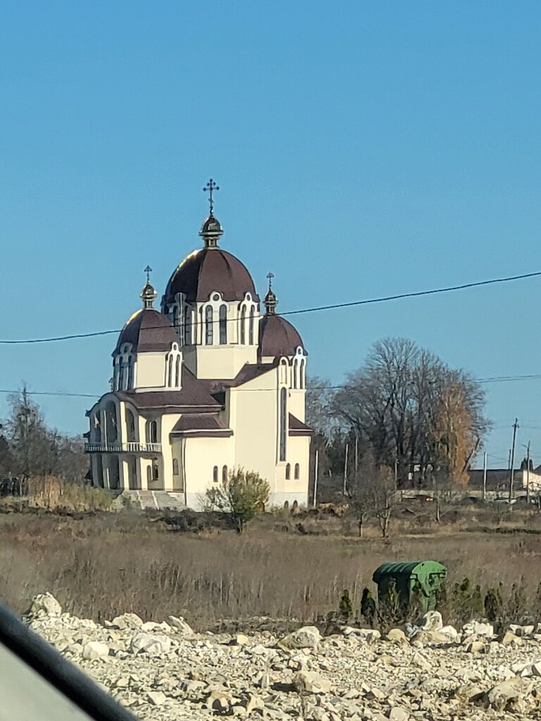 Kirche; ein weißes Gebäude, achteckige aneinandergesetzte Türme, ein paar rechteckige Elemente. Halbrunde dunkelbraune Kuppeln auf den Türmen.