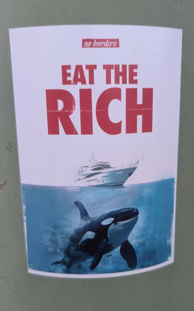 Ein Aufkleber auf einer grünen Laterne. Darauf der NO BORDERS  EAT THE RICH

Darunter eine Yacht und ein Orca 