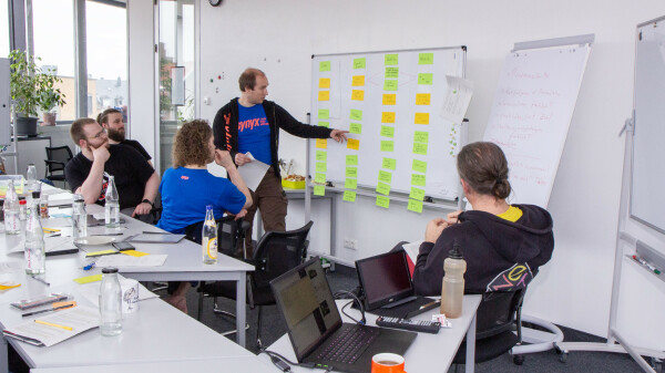 Auf dem Bild sind Eberhard Wolff und vier Teilnehmer zu sehen, die an einem Whiteboard in einem Büro diskutieren. Auf Tischen sind verschiedene Gegenstände wie Flaschen, Notebooks und Schreibmaterialien zu finden. Das Zimmer hat natürliches Licht durch Fenster im Hintergrund.
