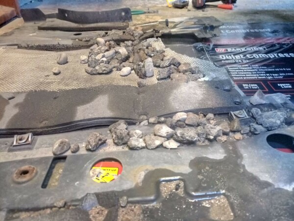 A garage floor with an under engine cover dropped down. There are large gravel rocks and chunks of asphalt on it, a lot of them.