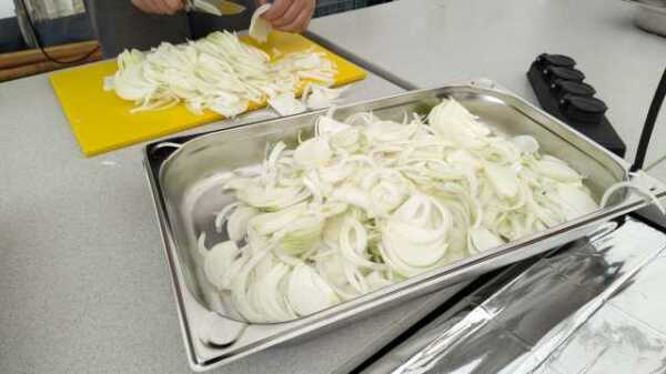 Ein Behälter voll geschnittener Zwiebeln auf einem weißen Tisch. Im Hintergrund sieht man ein gelbes Schneidebrett und die Hände einer Person, die gerade Zwiebeln schneidet.