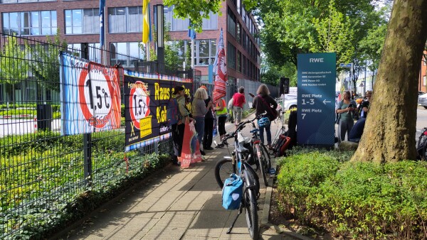 Gehweg mit darauf stehenden Fahrrädern und Menschen. Links ein Zaun mit Bannern, die das 1,5 Grad-Ziel thematisieren. Rechts das Firmenschild von RWE.