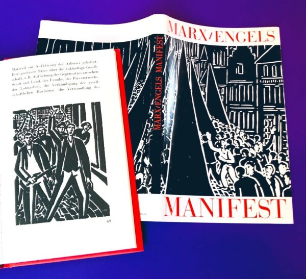 Neben dem Schutzumschlag mit Schwarz-Weiß-Holzschnitten von Frans Masereel liegt eine aufgeschlagene Seite mit einer weiteren Werk-Abbildung: Es handelt sich um die illustrierte Ausgabe vom »Manifest der Kommunistischen Partei« von Karl Marx und Friedrich Engels.