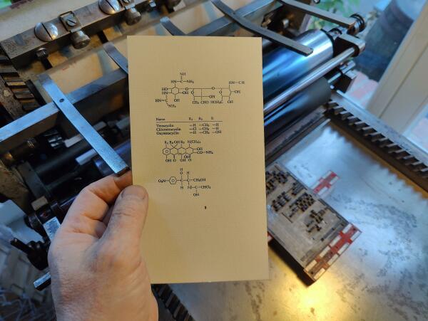 Foto des mit Satzmagneten auf dem Fundament einer kleinen Zylinderpresse, einer Korrex Modell Stuttgart, montierten Bleisatzes.  Davor von einer Hand gehalten der Abzug der Form auf Vorsatzpapier. Der Bleisatz besteht aus chemischen Formeln.