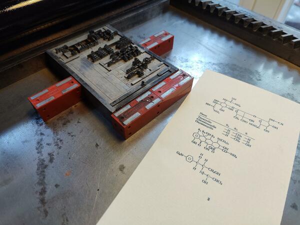 Foto des mit Satzmagneten auf dem Fundament einer kleinen Zylinderpresse, einer Korrex Modell Stuttgart, montierten Bleisatzes.  Daneben ein Abzug der Form auf Vorsatzpapier. Der Bleisatz besteht aus chemischen Formeln.