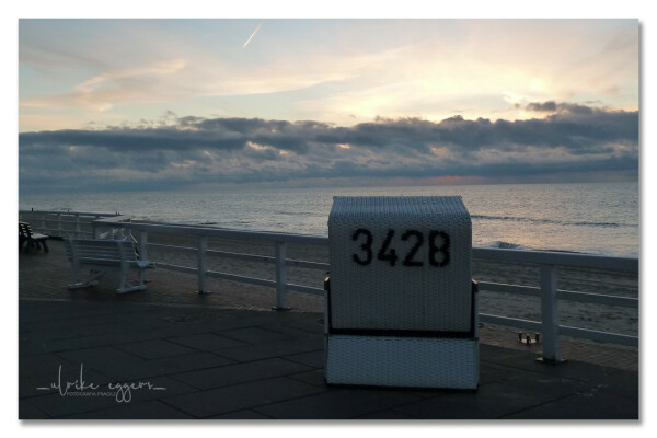 Blick von der etwas erhöhten Promenade in Westerland / Sylt auf die Nordsee im Abendlicht. Vorne steht ein weißer Strandkorb, auf seinem Rücken groß die Nummer 342B. Links davon zwei weiße Bänke, die Promenade ist mit einem weißen Holzgeländer gesichert.