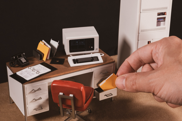 Miniature 1:12 scale model of a desk and chair. The desk is white with a wood desktop. On it from left to right is a black telephone, an open ring binder, a inbox shorter with files in it and a computer terminal. A minicomputer stands on the right, a normal sized hand is on the right taking a file folder from an open drawer.