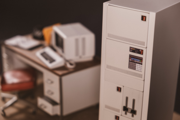 A miniature 1:12 scale diorama. In foreground on the right, a IBM Series\1 minicomputer from 1976. On the left, out of focus, a desk with all sort of things on it including computer terminal. 