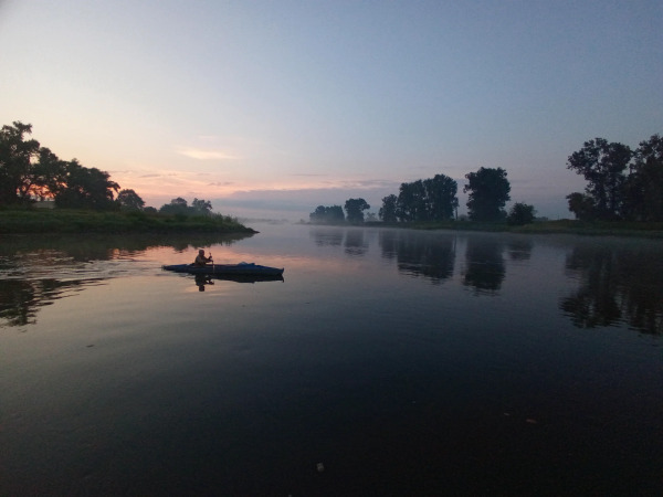 Ich stehe am Ufer der Elbe, dessen Wasser sich vor uns ausbreitet.
Von links geht die Sonne bald auf und man sieht schon die ersten reflexionen auf dem Wasser 
Über dem Fluss sieht man noch leichte Nebelschwaden über dem Wasser stehen.
Die Bäume am gegenüberliegenden Ufer spiegeln sich im leicht hellblau aussehenden Wasser

Genau vor uns paddelt gerade ein Wassersportler mit seinem Faltboot vorbei