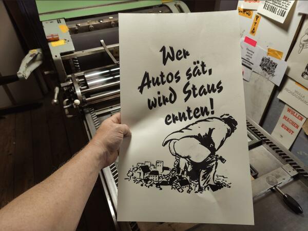 Foto einer Hand, die einen Probeabzug vor die Druckmaschine hält. Das Motiv ist ein Mann im Anzug, der eine Innenstadt mit Autos vollscheisst. Darüber in der 1940er Pinselschrift Reporter der Text: Wer Autos sät, wird Staus ernten!