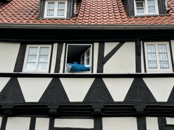 Ein Fachwerkhaus mit einem offenem Fenster aus dem ein bkaues Kissen guckt. Quasi zum Lüften herausgelegt. 