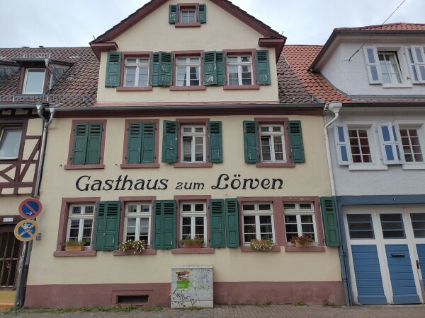 Foto einer Fassade eines Hauses. Es ist in gelb gehalten und hat grüne Fensterläden. In der Mitte steht Gasthaus zum Löwen. Das w sieht halb aus, wie ein n und halb wie ein w