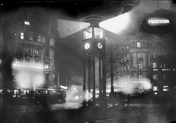 Fotografie zeigt den Verkehr am Potsdamer Platz in der Nacht. Mittig leuchtet der Verkehrsturm nach links und rechts. Die Autos erhellen die Straße. Rechts hinten die Leuchtreklame vom "Palast-Hotel". Nachweis von wiki commons folgt.