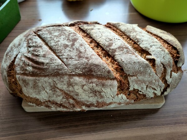 Dunkles Küchenbord im Fensterlicht auf dem ein Holzbrett mit einem Mischbrot drauf liegt.