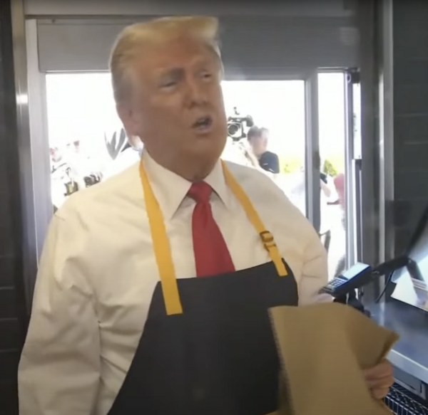 A picture of Donald Trump in a cramped McDonald's kitchen. He's wearing a white shirt, a red tie, and a cooking apron. He's talking a lot and holding a brown paper bag in his hand.