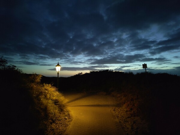 Gepflasterter Weg, links eine eckige Laterne mit hellem Licht. Rechts Dünen mit Bewuchs. Darüber der Himmel in der untergehenden Sonne mit einigen dunklen Wolken und viel blau. Im unteren Teil des Himmels noch ein Streifen orange.