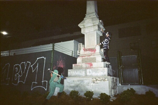 you can see two people in the dark and a monument covered in red (you can't tell which one). a hooded man with a green suit and a big hammer is hitting the monument on the lowest step, another man with a mask and a striped shirt is standing on the third step and spraying something on the statue. in the background you can see a black wall with a white graffiti "1 by 1",