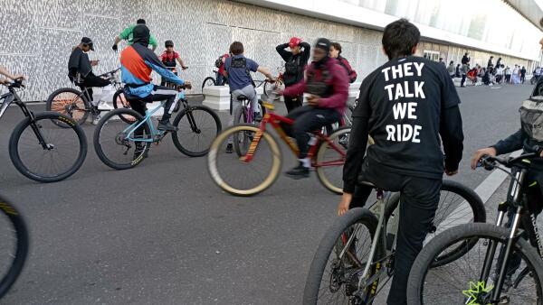 Junge Leute auf Fahrrädern, eine Person blickt weg von der Aufnahme Position und hat ein schwarzes Shirt mit der Aufschrift "They talk we ride"