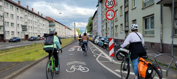 Radfahrende auf der Schloßstraße, auf der Fahrbahn ist das Verkehrszeichen für Überholverbot und ein Sharrow markiert. Die Türzone ist durchgehend mit vielen "X" markiert. Im Vordergrund hängen Verkehrszeichen für Tempo 30 und Überholverbot für einspurige Fahrzeuge