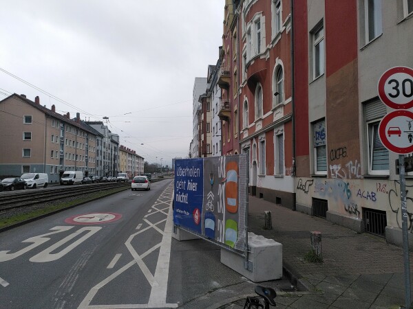 Blick in die Schloßstraße mit aufgestellten Schildern und auf der fahrbahn aufgemalten Piktogrammen für Tempo 30 und Überholverbot von Radfahrenden, dazu ein großes Plakat mit der Aufschrift "Überholen geht hier nicht!". Die Türzone der Parkplätze ist komplett mit x-en markiert