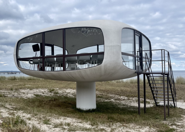 Farbiges Querformat. Ein Bauwerk mit großen Fenstern und einer Metalltreppe rechts steht auf einer Düne. Dahinter ein schmaler Streifen Meer, darüber Himmel und Wolken. Das Bauwerk ruht auf einem zentralen Pfeiler, besteht nur aus einem einzigen Raum mit Fenstern nach allen Seiten und ist vom Dach, Boden und Ecken her abgerundet. 